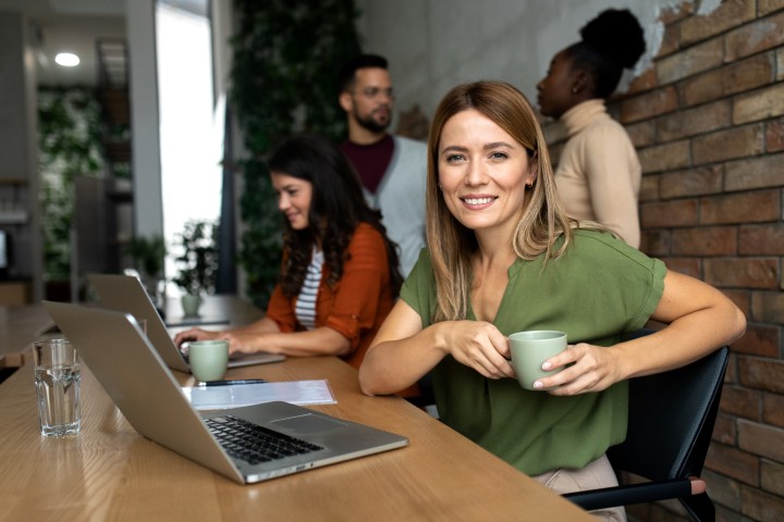 Frau mit langen blonden Haaren hält eine Tasse in den Händen und schaut lächelnd in die Kamera, im Hintergrund sitzt eine weitere Frau an einem Computer und dahinter sind unscharf weitere Kollegen zu sehen