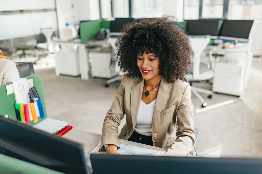 Eine junge, dunkeljahrige Frau mit Afro-Frisur sitzt in einem Büro am Schreibtisch und arbeitet mit zwei Bildschirmen an einem Rechner.