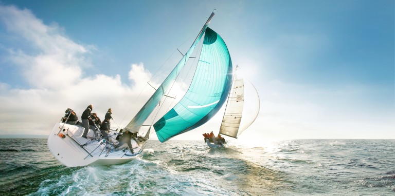 Segelboote bei einer Regatta auf offenem Meer mit gesetzten Segeln und mehreren Personen an Bord.