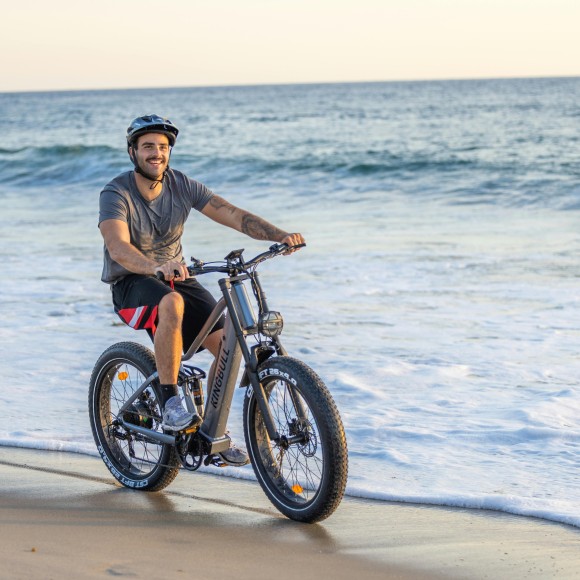 Junger Mann mit Helm fährt lachend auch einem E-Bike am Strand entlang