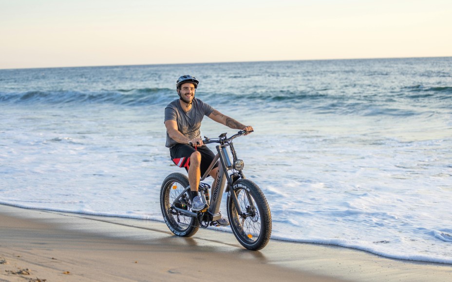 Junger Mann mit Helm fährt lachend auch einem E-Bike am Strand entlang