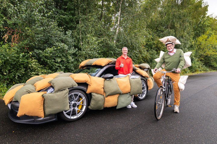 Ralf Schumacher steht in der Tür eines von Kissen ummantelten Autos. Neben ihm sitzt ein Mann mit Kissen am Körper auf einem Fahrrad.