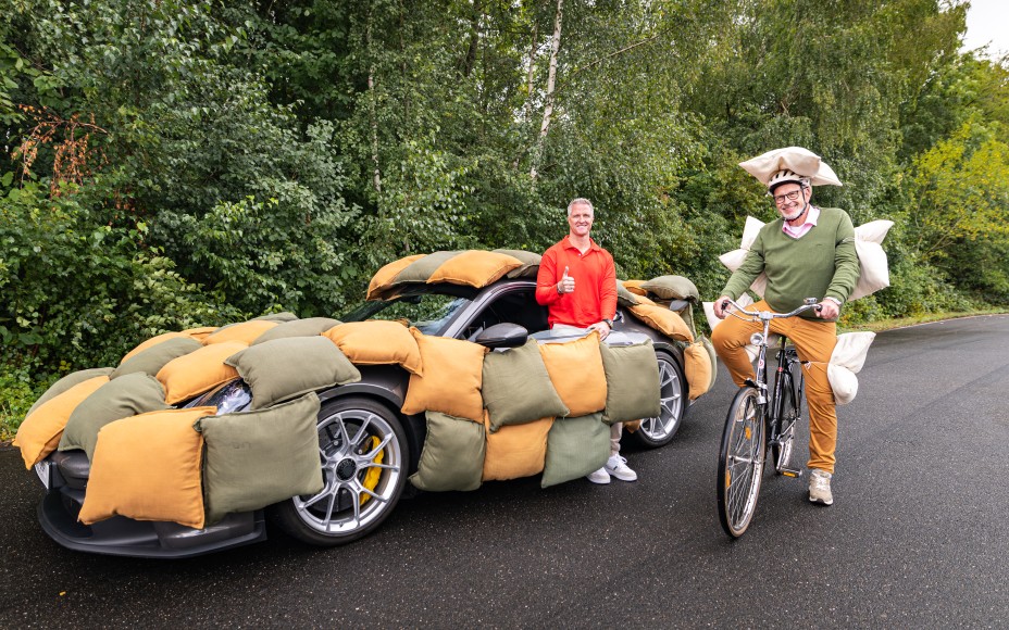 Ralf Schumacher steht in der Tür eines von Kissen ummantelten Autos. Neben ihm sitzt ein Mann mit Kissen am Körper auf einem Fahrrad.