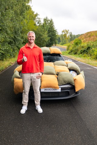 Ralf Schumacher steht vor einem mit Kissen ummantelten Auto und zeigt Daumen hoch.