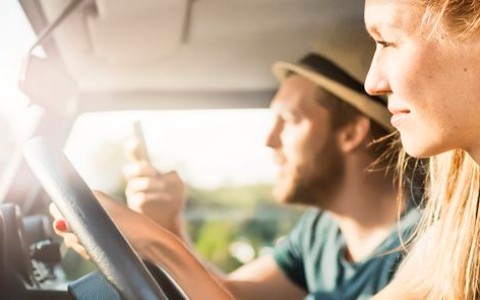 Junge blonde Frau fährt, während der ebenfalls junge Beifahrer mit Hut sein Handy hochhält und sie etwas zu suchen scheinen