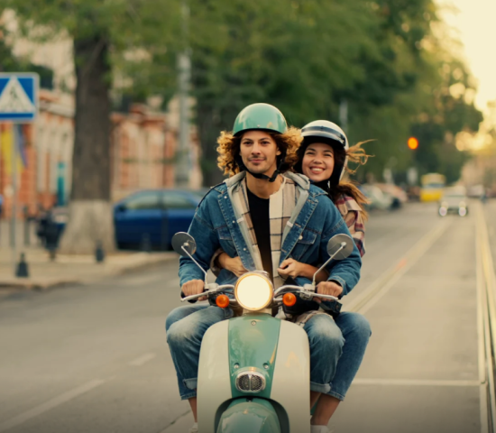 Junges Paar fährt gemeinsam mit Helmen auf einem Motorroller durch eine Stadt.
