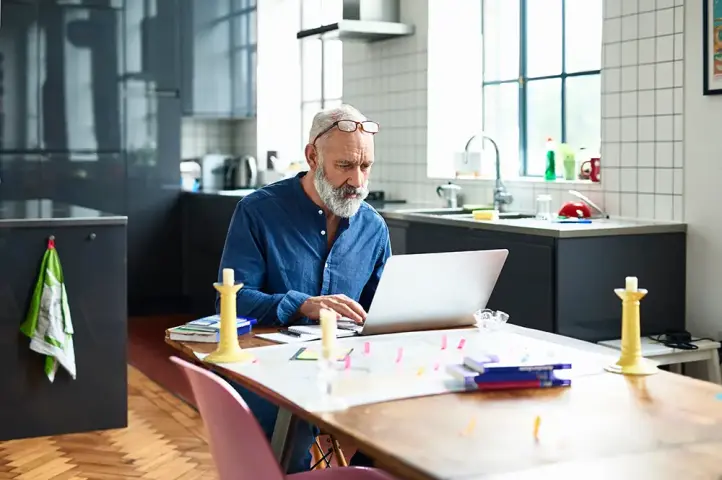 Älterer Mann arbeitet mit einem Laptop an einem Tisch in einer modernen Küche.