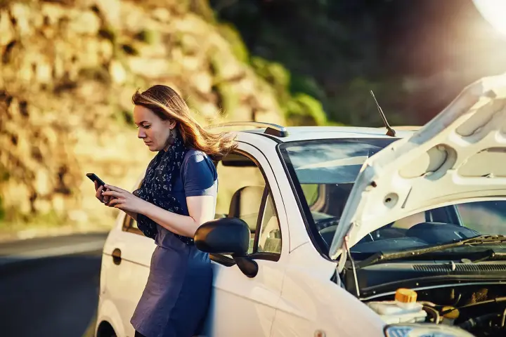 Frau mit Smartphone neben einem Auto mit geöffneter Motorhaube bei einer Panne.