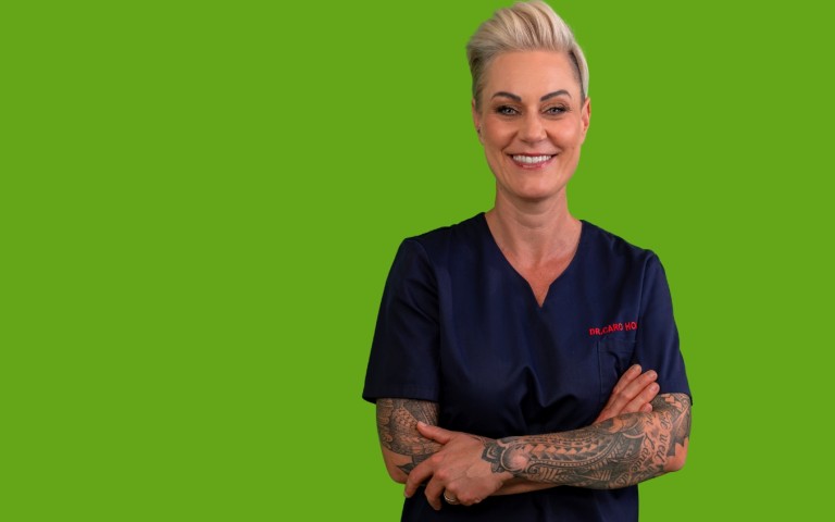 Doc Caro steht mit verschränkten Armen und dunkelblauem OP-Anzug vor HDI-grünem Hintergrund