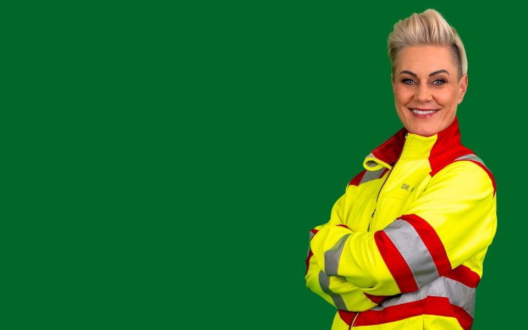 Doc Caro steht mit verschränkten Armen in einem gelb-roten Sanitäter-Anzug vor grünem Hintergrund