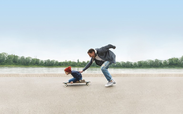 Vater schiebt auf Skateboard knienden Sohn an