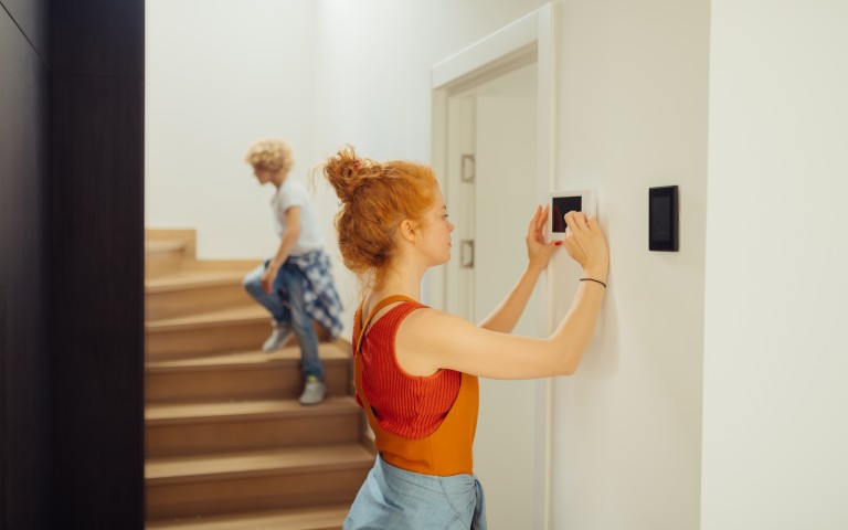 Frau mit roten, hochgesteckten Haaren im orangefarbenen Shirt programmiert eine Alarmanlage an der Wand, während im Hintergrund ein Kind eine moderne Holztreppe nach oben geht