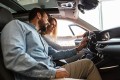 Junge blonde Frau sitzt am Steuer eines stehenden Autos und schaut auf die Armaturen, während ein Mann mit Dreitagebart ihr dort lächelnd etwas erklärt