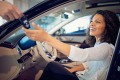 Frau mit dunklen Locken sitzt in einem Auto, vermutlich in einem Autohaus und nimmt einen Schlüssel aus einer männlichen Hand entgegen
