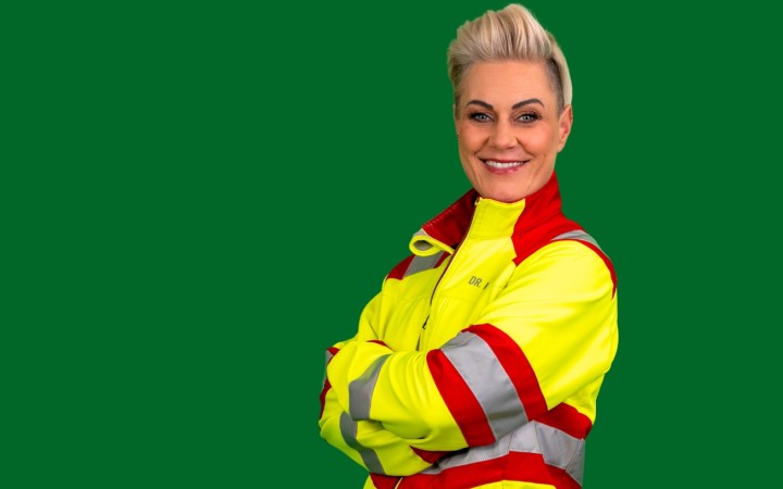 Doc Caro steht mit verschränkten Armen in einem gelb-roten Sanitäter-Anzug vor grünem Hintergrund