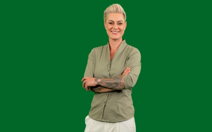 Doc Caro steht mit verschränkten Armen in sportlicher Kleidung vor grünem Hintergrund