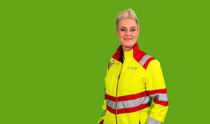Doc Caro steht in neongelber Rettungssanitäterjacke vor hellgrünem Hintergrund und läßt die Arme locker an der Seite herunterhängen