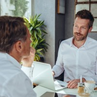 Zwei Männer in weißen Hemden sitzen sich während eines Beratungsgesprächs an einem Schreibtisch gegenüber