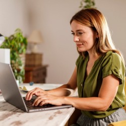 Junge Frau auf gelbem Polsterstuhl sitzt an einem Tisch an ihrem Laptop und tippt etwas