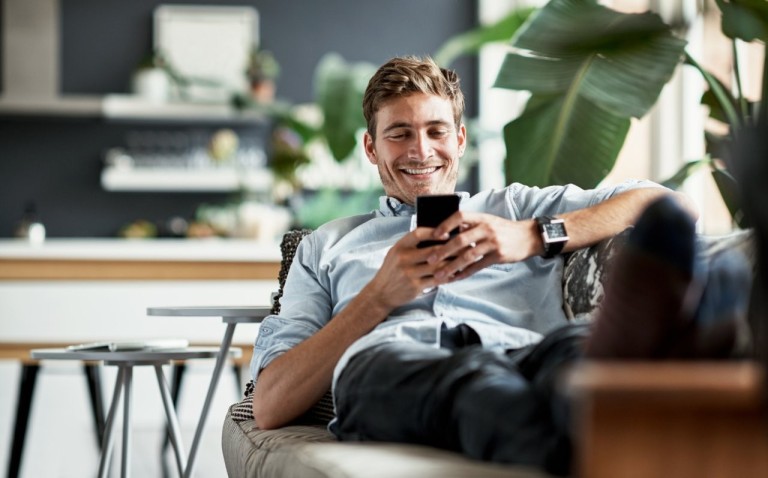Mann sitzt lächelnd auf dem Sofa und schaut auf sein Handy