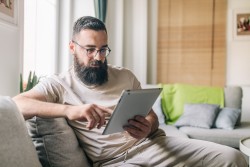 Mann mit langem dunklen Bart nutzt Tablet zuhause, um online Feedback oder eine Beschwerde bei der Versicherung einzureichen.