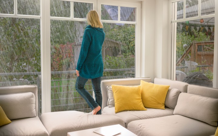 Frau mit langen blonden Haaren und grüner langer Strickjacke steht barfuß auf einem weißen Ecksofa mit gelben Kissen, das vor einer Fensterfront steht, und schaut nach draußen in ein Unwetter, beruhigt, da sie ein Elementarversicherung hat.