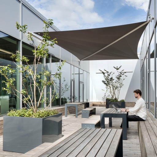 Mann sitzt in weißem Hemd auf einer modernen, begrünten Dachterrasse an einem Holztisch auf einer Holzbank und arbeitet an einem Laptop