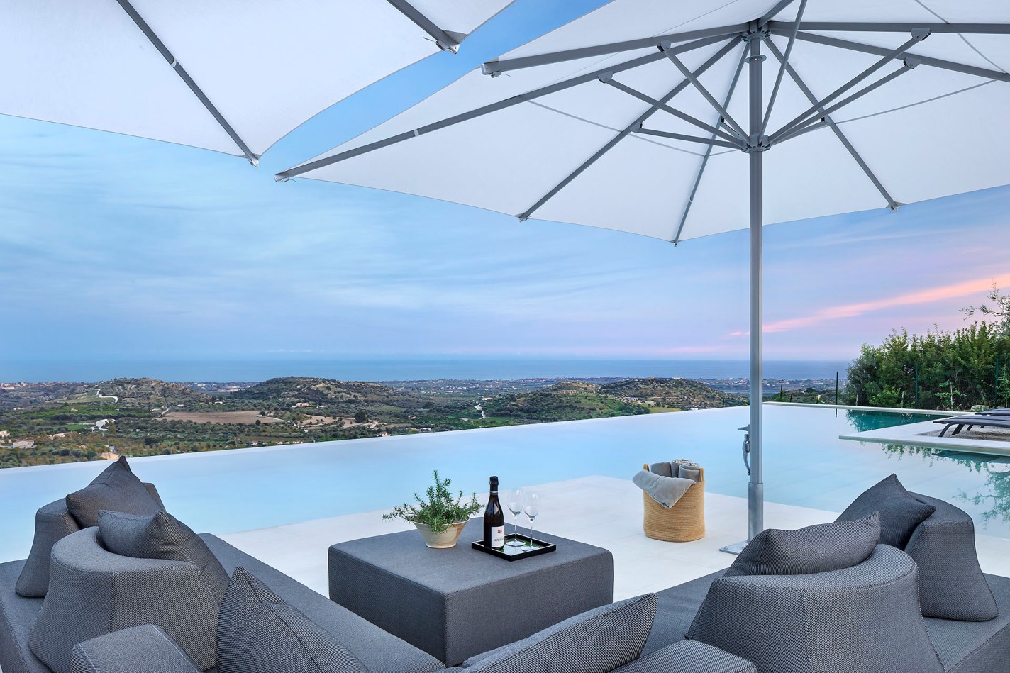 Terrasse mit grauen Loungemöbeln und weißen Sonnenschirmen am Infinity-Pool, mit Blick auf grüne Hügel und das Meer bei sanftem Abendlicht.