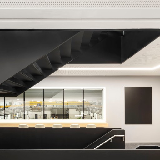 Sieben weiße Stühle stehen in einer Reihe an einem hellen Konferenztisch aus Holz, der vor einer großen Glasfront steht, durch die man in eine große moderne Produktionshalle blickt