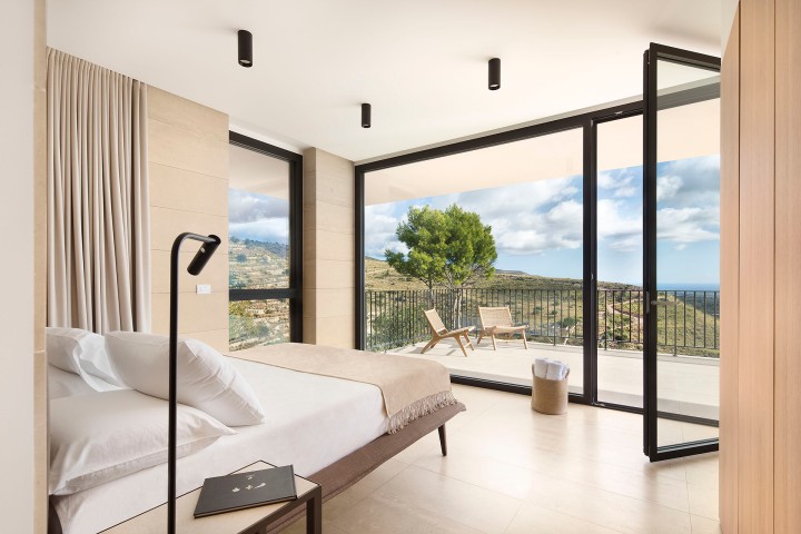 Helles, modernes Schlafzimmer mit Panoramafenster und Blick auf eine Terrasse mit Liegestühlen und hügeliger Landschaft.