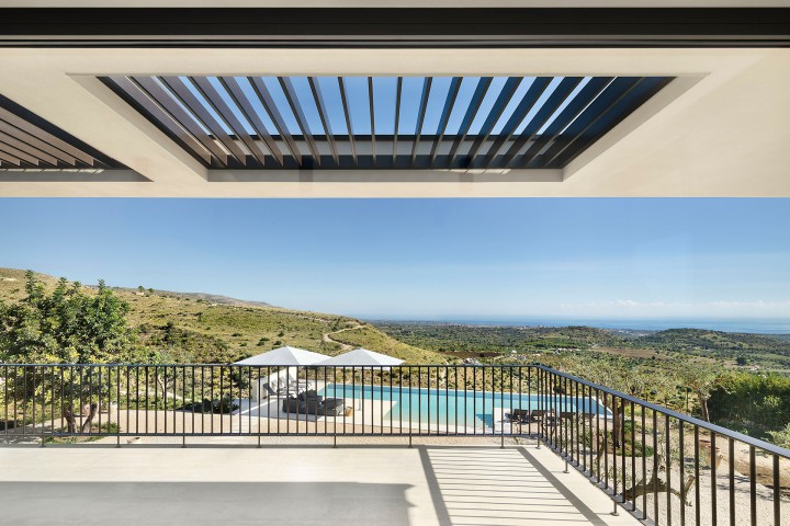Balkon mit modernem Geländer und Blick auf Pool, Sonnenschirme und grüne Hügel bis zum Meer unter klarem blauem Himmel.