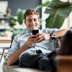 Mann sitzt lächelnd auf dem Sofa und schaut auf sein Handy