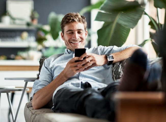 Mann sitzt lächelnd auf dem Sofa und schaut auf sein Handy
