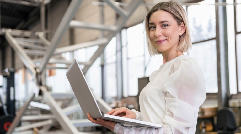 Blonde Frau mit kinnlangen Haaren und weißer Bluse steht mit einem Laptop in den Händen in einer Betriebshalle und lächelt in die Kamera.