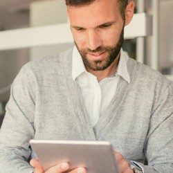 Mann in grauem Pullover steht in einem Büro und schaut etws auf einem Tablet
