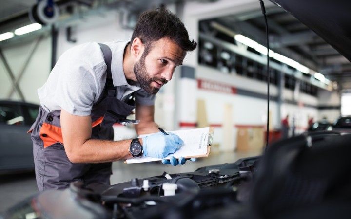 Mechaniker mit grauer Latzhose beugt sdich in den Motorraum eines Autos, das in einer Werkstatt steht und inspiziert etwas