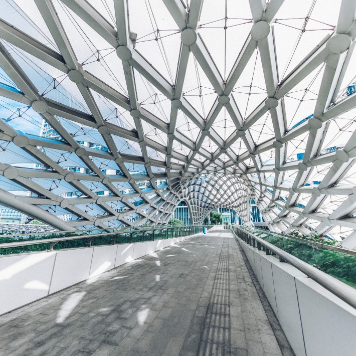 Innenansicht einer modernen, architektonischen Fußgängerbrücke mit einem auffälligen, wellenförmigen Dach aus weißen Stahlträgern und Netzen, das das Tageslicht filtert.