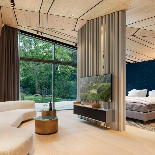 Modernes Hotelzimmer in hellen Beige- und Brauntönen, helles Holz an Wänden, Decke und Böden, cremefarbenes futuristisches Sofa, Flatscreen an einem Raumtrenner, Blick durch eine große Fenterfront in einen Garten