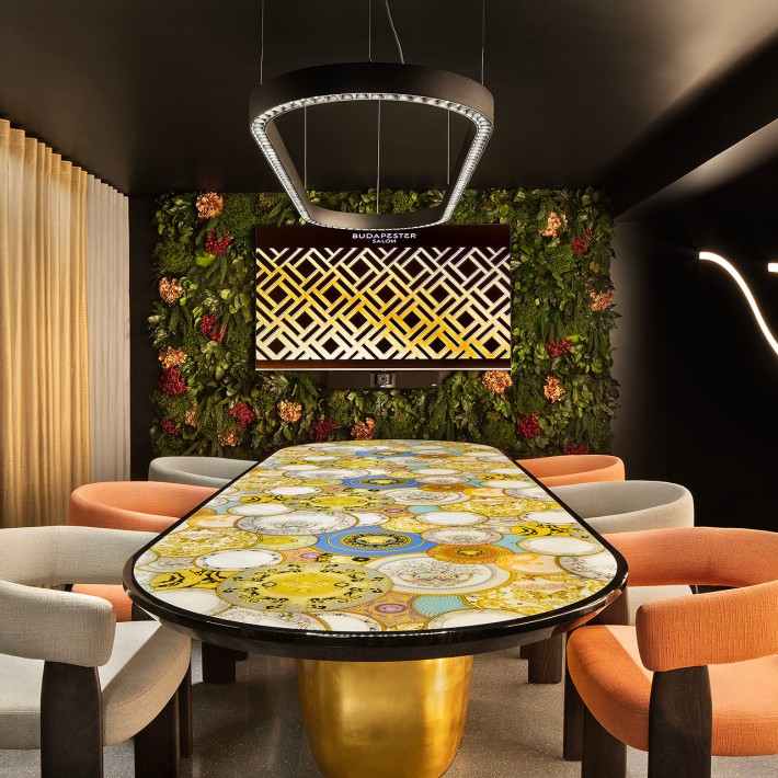 Innenansicht Budapester Salon mit einem großen Tisch mit großflächigem Mosaik auf der Platte, goldenem Fuß, darum Sessel in Naturtönen, die linke Wand besteht aus cremfarbenen Faltenvorhängen, die Kopfwand ist eine Blumentapete in dunklen Tönen und die Decke ist schwarzt