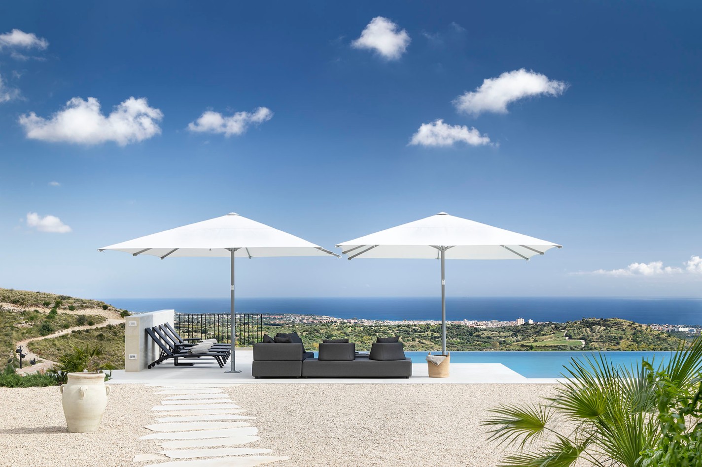 Moderne Terrasse mit Deko-Amphoren, einem rechteckigen Pool neben einer grauen Lounge-Landschaft unter zwei weißen Sonnenschirmen, im Hntergrund sonnige Hügel und das Meer