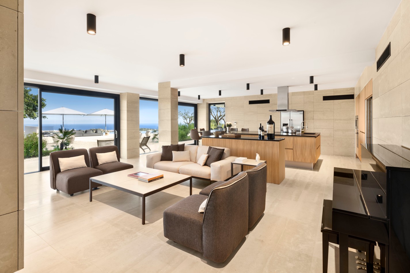 Moderne offene Küche mit Wohnbereich und Essecke in hellen Naturtönen, in der Küche stehen große Weinflaschen, im Hintergrund ist eine große Fensterfront mit Blick auf die Terrasse mit Sonnenschirm und Meer in weiter Ferne