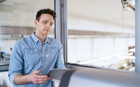 Mann im Jeanshemd lehnt sich entspannt an einen hohen Tisch und schaut etwas auf einem Tablet, im Hintergrund ist ein Büro zu sehen