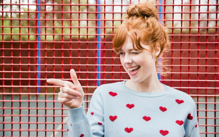 Rothaarige Frau mit hohem Dutt in hellgrauem Wollpullover mit roten Herzen zeigt schelmisch lachend auf jemanden während sie vor einem roten Metallgitter steht