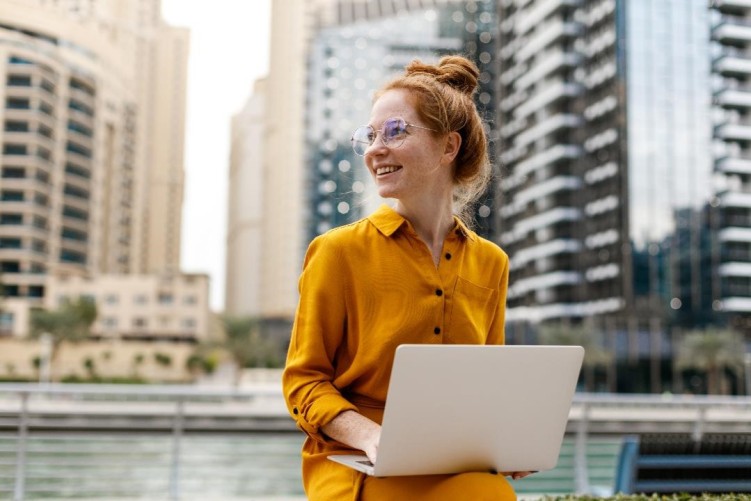 Junge Frau mit Brille sitzt in Dubai Marina Arie lächelnd am Laptop