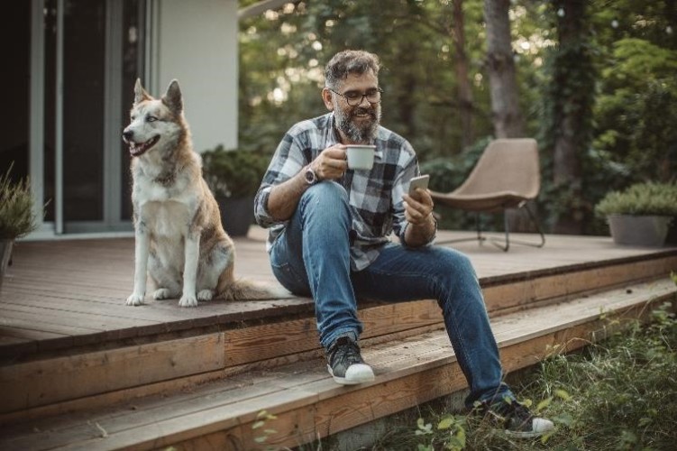 Mann sitzt mit Kaffee und Smartphone neben seinem Hund auf der Terasse