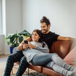 Junges Paar kuschelt auf dem Sofa während sie gemeinsam auf das Smartphone der Frau schauen