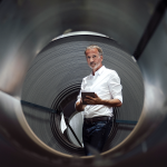 Mann mit weißem Hemd und grauem Bart steht mit einem Tablet in der Hand vor einer schwarzen Röhre