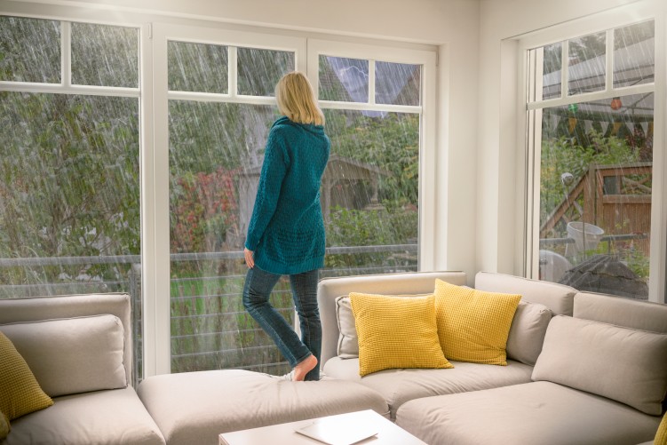 Frau mit langen blonden Haaren und grüner langer Strickjacke steht barfuß auf einem weißen Ecksofa mit gelben Kissen, das vor einer Fensterfront steht, und schaut nach draußen in ein Unwetter, beruhigt, da sie ein Elementarversicherung hat.