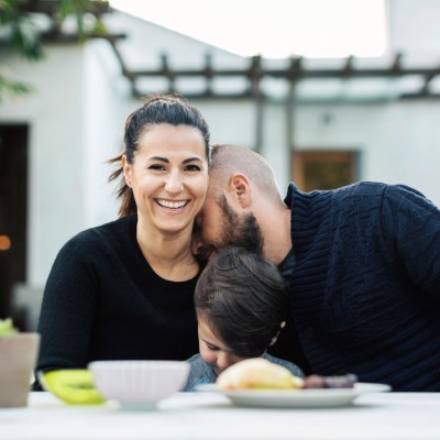 Familie sitzt vor ihrem Haus im Garten an einem Tisch, Frau lacht in die Kamera und Mann lehnt Kopf auf ihre Schulter