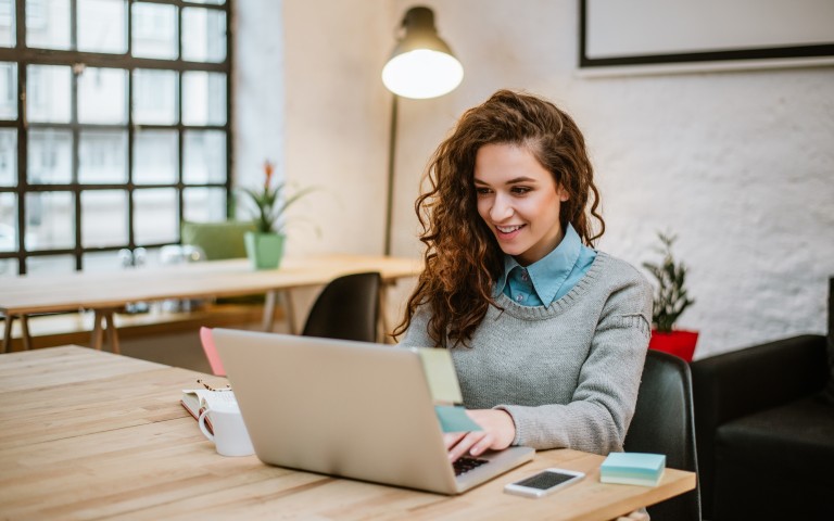 Eine junge Frau sitzt an einem Tisch und arbeitet am Laptop.